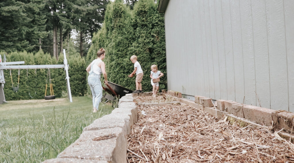 Christian homemaker wearing a white tank top and denim overalls and her two young children work together in a garden bed, with the children taking bricks from a wheelbarrow and placing them next to the house.